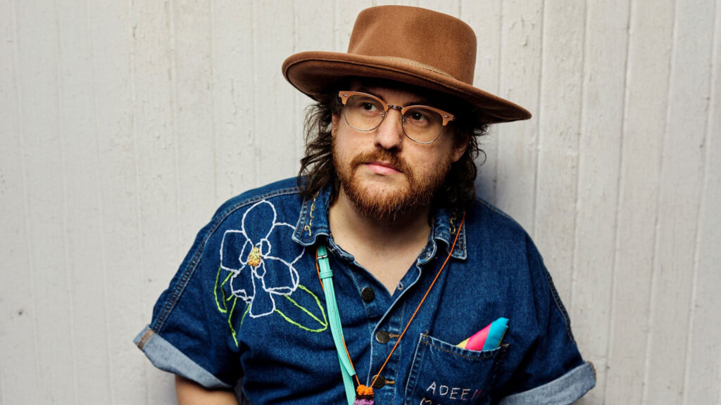 Promotional portrait of country-folk singer-songwriter Adeem the Artist wearing a brown brimmed hat, glasses, and a embroidered denim shirt.